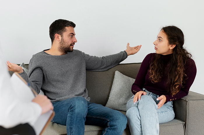 Couple in a heated discussion on the sofa, man gestures widely while woman looks concerned.