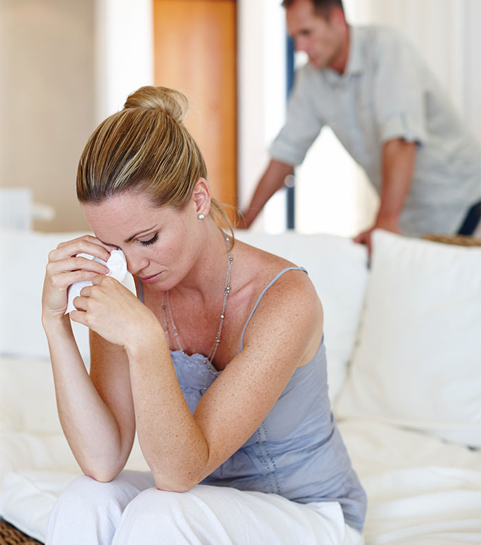 Woman sitting on couch tearfully holding tissue, feeling guilty about late father’s estate while man stands in background.