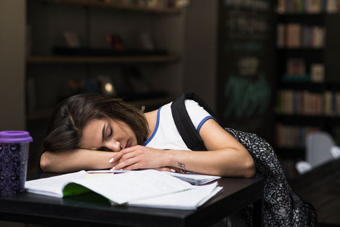 Student sleeping on books in library, tired from weeks of spiked drinks and sleep aids by roommate conflict.
