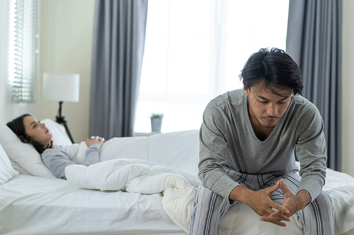 Woman upset and lying in bed with injured leg, fiancé sitting beside her looking worried after accident consequences.
