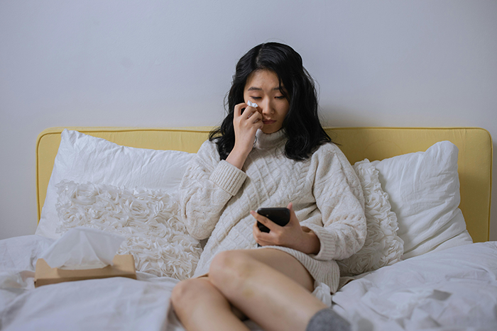 Woman on bed choosing affordable getaway with husband, looking at phone and wiping tears, facing criticism from bride.