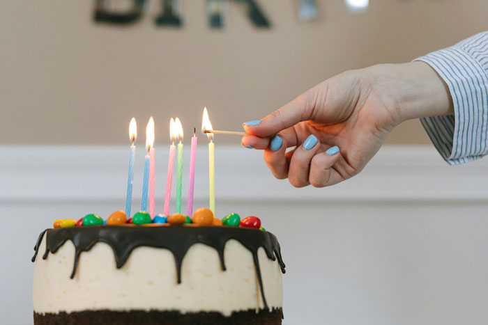Hand with blue nail polish lighting candles on birthday cake with colorful candies and chocolate drip frosting.